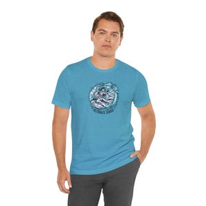 Op de afbeelding: Een lichtblauw t-shirt met een cirkelvormige afbeelding van een figuur in water en de tekst "Ultimate Surge". Het shirt is gemaakt van zacht materiaal en heeft korte mouwen.