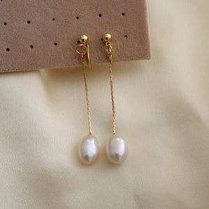 Peut inclure: Une paire de boucles d'oreilles pendantes dorées avec des perles blanches de forme ovale. Les boucles d'oreilles sont suspendues à une petite boule dorée et à une fine chaîne dorée. Le fond est beige clair.