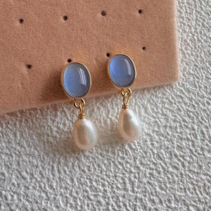 Puede incluir: Un par de pendientes dorados con cabujones ovalados de color azul claro y perlas blancas en forma de lágrima. Los pendientes se exhiben sobre una superficie rosa con pequeños puntos negros, mostrando un diseño de joyería delicado y elegante.