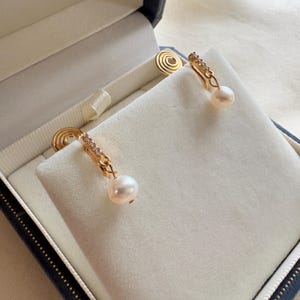 Peut inclure: Une paire de boucles d'oreilles dorées avec une perle en goutte et de petites pierres transparentes. Les boucles d'oreilles sont présentées dans un écrin à bijoux bleu et blanc avec un intérieur en velours. Design élégant.