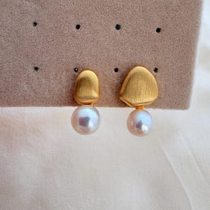 Peut inclure: Une paire de boucles d'oreilles composée de formes asymétriques en métal doré brossé fixées à des perles blanches rondes. Les boucles d'oreilles sont présentées sur un support en tissu beige perforé, mettant en valeur le contraste entre la finition dorée mate et la surface lisse et lustrée des perles.