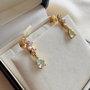 Peut inclure: Boucles d'oreilles dorées avec un clou en spirale et un design pendant. Chaque boucle d'oreille présente un groupe de petites pierres transparentes et une pierre précieuse en forme de poire, vert pâle. Les boucles d'oreilles sont présentées dans un écrin bleu avec un intérieur blanc.