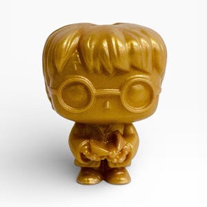 May include: A gold-colored figurine of a character with round glasses and a small book. The figure has a stylized design with a large head and small body. The figurine is standing upright against a white background.