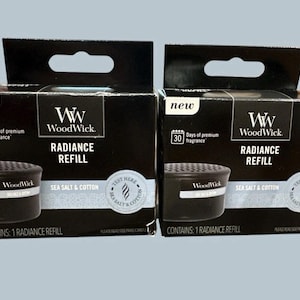 Puede incluir: Dos cajas negras de recarga WoodWick Radiance. Cada caja tiene una asa y presenta el texto "Sea Salt & Cotton". Las cajas también muestran "30 días de fragancia premium" y "Contiene: 1 Radiance Refill".