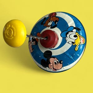 May include: Vintage toy top with a yellow handle and a blue and white spinning disc featuring Mickey Mouse, Donald Duck, and Goofy. The top has a red center and a silver metal rod.