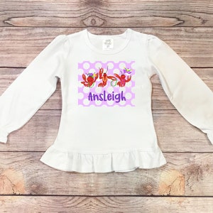 May include: White long-sleeved shirt with a purple and white polka dot design featuring three cartoon crawfish wearing Mardi Gras beads and a purple mask. The shirt has a ruffled hem and the name "Ansleigh" is printed below the crawfish.