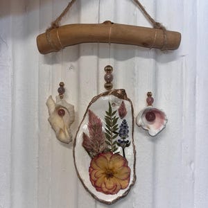 May include: A handcrafted mobile featuring a driftwood bar and hanging seashells. The mobile includes an oyster shell decorated with dried flowers and greenery, and is accented with pink beads. The seashells are suspended by thin wire.