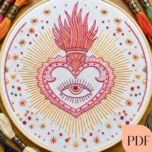 May include: Embroidered heart design in a wooden hoop. The heart is pink with an eye in the center, surrounded by radiating gold lines and starbursts. The top of the heart has a flame design in shades of orange and pink. Embroidery floss in various colors is visible.