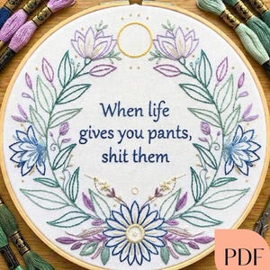 Op de afbeelding: Geborduurde borduurring met de tekst "When life gives you pants, shit them." Het ontwerp bevat bloemelementen in blauw, paars en groen. Borduurgaren in verschillende kleuren omringen de ring. Een PDF-pictogram staat rechtsonder.