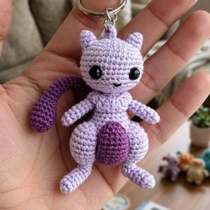 May include: A handmade crochet keychain featuring a purple Mewtwo character. The keychain has a silver ring and a small, smiling Mewtwo figure with black eyes, a purple tail, and a darker purple belly. The figure is made of soft yarn.