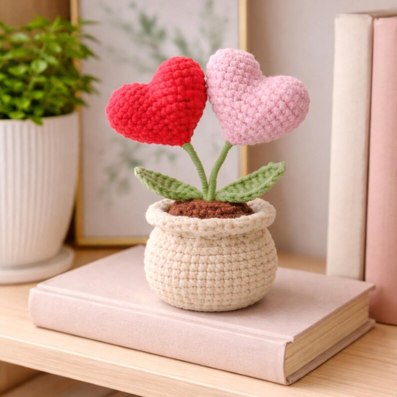 May include: A crocheted flower arrangement featuring two heart-shaped blooms, one red and one pink, in a beige pot. The stems are green, with green leaves. The pot sits on a light pink book, suggesting a decorative or gift item.