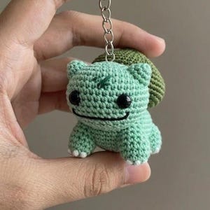 Könnte beinhalten: Ein gehäkelter Bulbasaur-Schlüsselanhänger in Hellgrün mit schwarzen Augen und einem lächelnden Mund. Der Schlüsselanhänger hat eine grüne Knolle auf dem Rücken und eine silberne Kette. Der handgefertigte Artikel wird in der Hand einer Person gehalten.