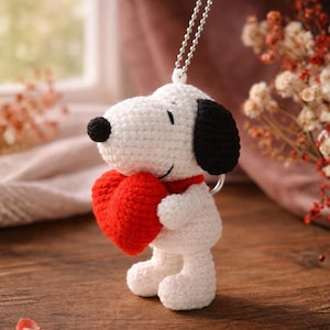 May include: A white and black crocheted Snoopy figure holding a red heart. The figure has a black nose and ears, a red collar, and a silver chain for hanging. The Snoopy figure is standing on a wooden surface.