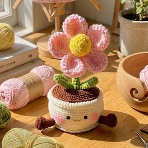 May include: A handmade crochet flower in a pot. The flower has pink petals and a yellow center. The pot is white with a smiling face and brown soil. Green leaves and brown arms complete the design. The flower is sitting on a wooden surface.