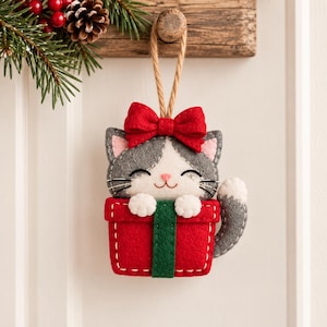 May include: A felt Christmas ornament shaped like a grey and white cat peeking out of a red gift box with a green ribbon. The cat has a red bow on its head, pink inner ears, and stitched details. A tan cord is attached for hanging the ornament.