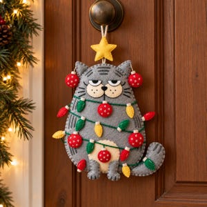 May include: A grey felt cat ornament hanging from a door knob. The cat has a grumpy expression, a yellow felt star on its head, and is wrapped in a string of colourful felt Christmas lights and red ornaments with white snowflake patterns.