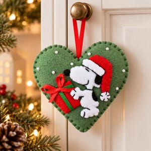 May include: A heart-shaped felt ornament in olive green featuring a white Snoopy character wearing a red Santa hat and holding a red gift box with a green ribbon. The ornament is accented with small white beads and hangs from a red ribbon loop.