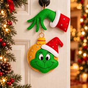 May include: A felt hanging decoration featuring a green hand with a red cuff and a yellow ornament shape with a green face and a red Santa hat. The hand and ornament are connected by a gold cord, with white star accents on the red fabric pieces.