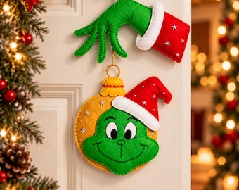 May include: A felt hanging decoration featuring a green hand with a red cuff and a yellow ornament shape with a green face and a red Santa hat. The hand and ornament are connected by a gold cord, with white star accents on the red fabric pieces.