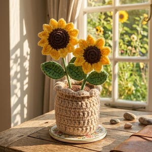 Pode incluir: Um arranjo de girassóis de crochê em um vaso bege. Os girassóis têm pétalas amarelas e centros marrons, com folhas e caules verdes. O vaso está sobre um descanso de copo com estampa floral, colocado em uma mesa de madeira perto de uma janela.