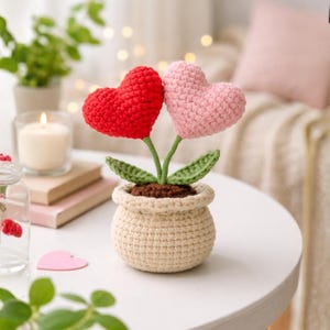 May include: A crocheted heart-shaped flower arrangement in a beige pot. The arrangement features a red and a pink heart, green stems and leaves, and brown soil. The pot is sitting on a white surface.