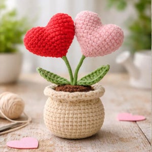 May include: A crocheted heart-shaped flower arrangement in a beige pot. The arrangement features a red and a pink heart, green stems and leaves, and brown soil. The pot is also crocheted.