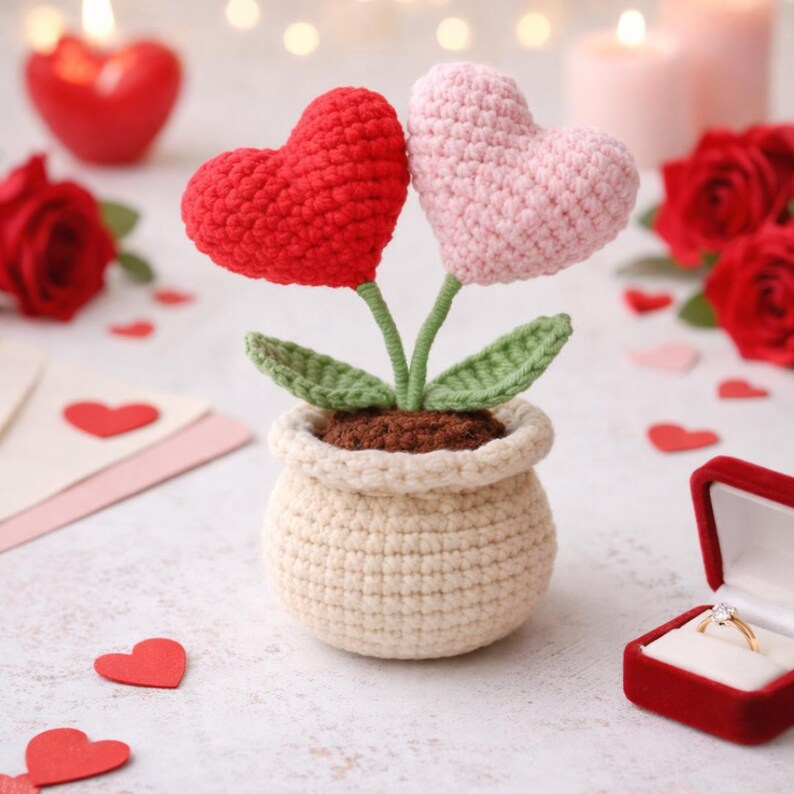 May include: A crocheted plant with two heart-shaped flowers, one red and one pink, in a beige pot. Green leaves and stems support the hearts. The background includes a red velvet ring box with a diamond ring, red roses, and heart-shaped confetti.