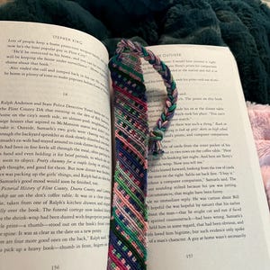 May include: A colorful, handmade bookmark with a braided top and a patterned design in shades of blue, pink, green, and purple. The bookmark is placed in an open book.