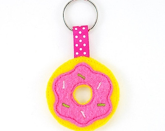 Può includere: Un portachiavi a forma di ciambella in feltro rosa e giallo con codette bianche. La ciambella ha un buco al centro ed è attaccata a un nastro rosa con pois bianchi.