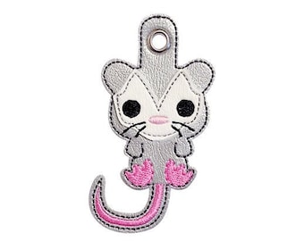 May include: A gray and pink embroidered opossum keychain with a loop for attaching to a bag or keys.
