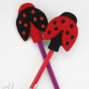 May include: Two red and black ladybug pencil toppers with black felt wings and red felt bodies. The ladybugs have black dots on their wings. The ladybugs are on two pencils, one purple and one red.