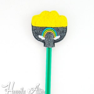 May include: A yellow and gray felt pencil topper with a rainbow and clouds design. The topper is attached to a green pencil.