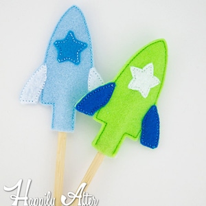 May include: Two felt rocket finger puppets on wooden sticks. One rocket is blue with a blue star and the other is green with a white star. Both rockets have blue wings.