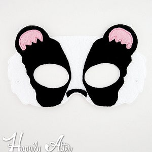 May include: A black and white felt mask with pink ears. The mask has eye holes and is shaped like a badger.