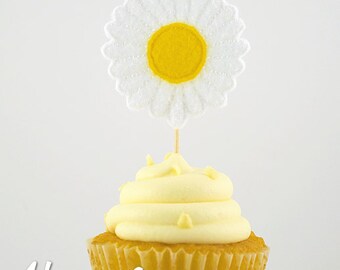 May include: A yellow cupcake with white frosting topped with a white daisy with a yellow centre. The daisy is on a toothpick.