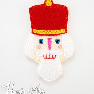 May include: A felt appliqué of a Nutcracker's face. The Nutcracker has a red hat with a yellow trim, a white beard, and a pink nose. The eyes are blue and the mouth is red.