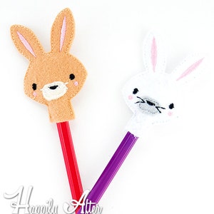May include: Two felt bunny pencil toppers. One is brown with pink ears and a red pencil. The other is white with pink ears and a purple pencil. The text "Happily After Designs" is at the bottom of the image.
