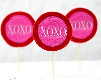 Könnte beinhalten: Drei rote und rosafarbene Filzkreise mit weißer Stickerei, auf denen jeweils "XOXO" steht. Die Kreise sind an Holzspießen befestigt.