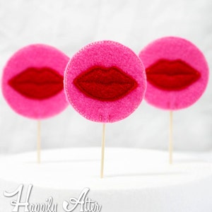 以下が含まれることがあります： 木製の串に赤いフェルトの唇がついたピンクのフェルトの円が3つ。円は縁に白いステッチで飾られています。