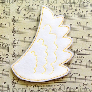 May include: A white felt applique with a yellow stitched outline in the shape of a wing. The applique is on a sheet of music.