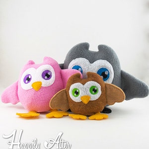 Puede incluir: Tres juguetes de peluche de búho, uno rosa, uno marrón y uno gris, con ojos y picos bordados. Los búhos están sentados sobre una superficie blanca. Happily After Designs está escrito debajo de los búhos.