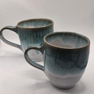 Peut inclure: Deux tasses en céramique avec un émail réactif présentant un dégradé de bleu sarcelle profond vers une base blanche. Chaque tasse possède une anse incurvée et un bord arrondi lisse, avec une finition brillante et des nuances subtiles.