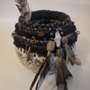 Può includere: Una ciotola rotonda in ceramica con base maculata verde e bianca, sormontata da un bordo rivestito in tessuto scuro. Il bordo è decorato con frange nere, perline di legno, piccole pietre grigie e diverse piume a righe grigie e bianche.