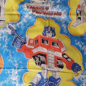 May include: A blue fabric sheet featuring a repeating pattern of Transformers characters including Optimus Prime, yellow sports cars, and robots. The print includes the Transformers logo and the text 1984 Hasbro Industries Inc. All Rights Reserved.