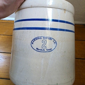 Puede incluir: Una vasija de cerámica cilíndrica de color blanco roto con dos rayas azules horizontales cerca del borde superior. El frente muestra un sello ovalado azul con el texto MARSHALL POTTERY CO., el número 2 y Marshall, Texas. La superficie tiene un esmalte craquelado.