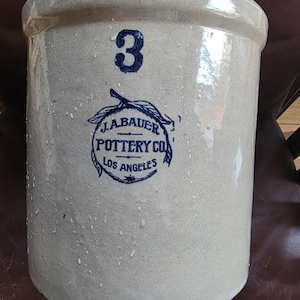 Puede incluir: Una vasija de gres beige de forma cilíndrica con un número 3 azul prominente en la parte superior. Debajo del número hay un sello circular azul con un diseño de hoja y el texto J.A. BAUER POTTERY CO LOS ANGELES sobre la superficie vidriada.
