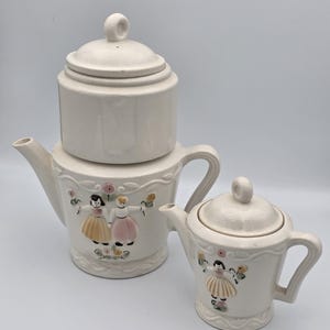 Puede incluir: Una tetera de cerámica color crema y una jarra de leche más pequeña a juego, ambas con figuras en relieve de personas con faldas sosteniendo flores. Cada pieza tiene una tapa con un asa circular, un pico curvo y un asa lateral decorada.