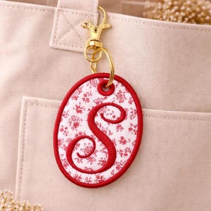 May include: Oval-shaped keychain with a red floral pattern and a red embroidered letter "S". The keychain is attached to a gold-colored clasp and ring, hanging against a beige tote bag.
