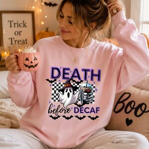 以下が含まれることがあります： 「DEATH before DECAF」の文字がプリントされたライトピンクのクルーネックトレーナー。魔女の帽子をかぶった白い幽霊、市松模様、ドクロのアイコンが描かれたコーヒーカップを持つ骸骨の手がデザインされています。