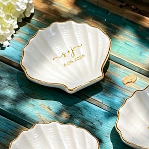 May include: Three white ceramic shell-shaped trinket dishes with gold-painted edges. Each dish features gold cursive lettering: one displays s + j and 6.06.2026, another shows v + a, and the third features g + b, all centered on the ribbed surfaces.